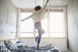 attractive woman jumping on bed in the morning, curly hair, casual style, blue jeans, white sweater, happy, natural window light