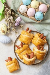 Traditional Easter symbols and food. Buns in the form of an Easter rabbit from sausage and cheese in a yeast dough with colored eggs on a gray stone background. View from above.