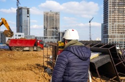Photo of a construction worker in a white helmet who looks into the level at the construction site of multi-storey buildings with a red truck on the street in the city.