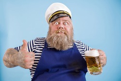 Joyful bearded person with overweight in sailor suit holds mug of tasty beer and shows thumb up on light blue background in studio