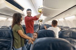 Cheerful man looking at his wife with a smile and putting carry on luggage in compartment while traveling together with his family by plane. Family vacation, transportation concept