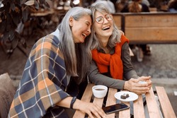Pretty senior Asian woman puts head on laughing friend shoulder resting together at table in street cafe on nice autumn day