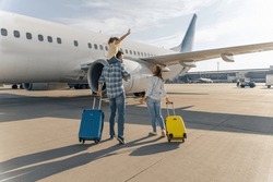 Back view of happy family standing near a large plane with two suitcases outdoor. Trip concept