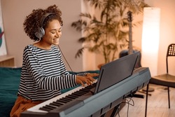 Joyful multiracial woman in headphones sitting on bed and smiling while playing melody on electronic musical instrument