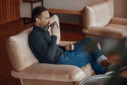Side view of adult man in cosy armchair drinking coffee while looking out the window
