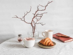 Cozy morning - cappuccino, croissant, vase with branches, leather planner notebook on the table in a bright cozy room. Cozy home interior       