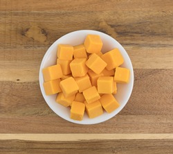 Top view of cubed mild cheddar cheese in a white bowl atop an old wood counter top.