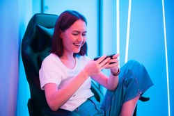 Cute gamer girl sit on a gaming chair, smiling and playing mobile online game on a smartphone. Having fun playing with friends online.
