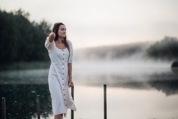 Tender romanitc sentimental female lady in the morning on a wooden pier near the misty river in a white dress