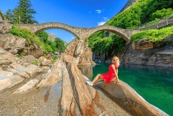 Woman resting sitting under the Ponte dei Salti, old Roman stone bridge. Verzasca River in Verzasca valley by Lavertezzo town. Riverside leisure and high diving in Ticino canton of Switzerland.