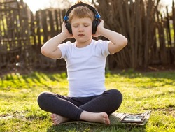 A red-haired teenager boy with headphones and closed eyes in a white T-shirt and gray pants listening to music on a mobile tablet and relaxing on the grass in the fresh air in nature