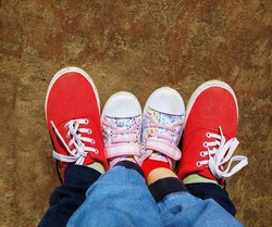 Top view of two pairs of legs, four feet, an adult and a child, shod in red and colored sneakers on a wooden surface
