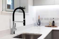 An elegant modern kitchen tap which is partly stainless steel and partly black in design with white quartz worktop and under mounted sink with a blurry background