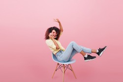 Funny girl in black sneakers and white socks posing in studio with pink interior and listening favorite song. Indoor photo of charming african woman in trendy jeans sitting on chair.