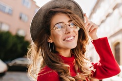 Close-up portrait of cheerful white woman in glasses touching her hat on blur background. Photo of fashionable girl with beautiful brown hair smiling to camera.