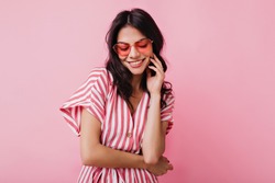 Romantic brown-haired girl in trendy heart glasses posing with shy smile. Indoor photo of graceful young woman in summer attire and accessories.