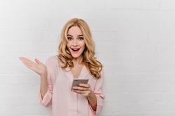 Adorable girl with phone in hand expressing positive emotions. Attractive fair-haired woman in cotton pink sleepwear holding smartphone near bricked wall.