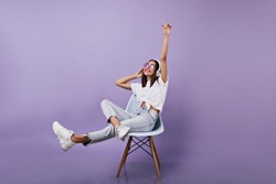 Joyful brunette girl listening music in studio. Relaxed female model in jeans posing on purple background with headphones.