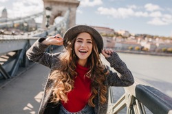 Gorgeous smiling woman with long curly hair posing with pleasure on bridge on blur background. Outdoor photo of blissful white lady enjoying autumn weekend in Europe.