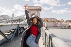 Shapely blissful girl in red sweater dancing on bridge on blur city background in autumn morning. Outdoor photo of happy female tourist having fun, exploring attractions.