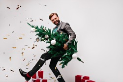 Blissful dark-haired man fooling around while preparing for new year. Laughing male model holding green christmas tree.