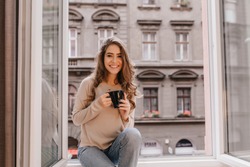 Adorable woman expressing positive emotions while posing on sill with cup of latte. Indoor photo of stunning female model with dark hair wears jeans and sitting beside window.