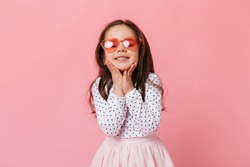 Close-up portrait of dark-haired little girl in polka-dot blouse and pink glasses, smiling on isolated background