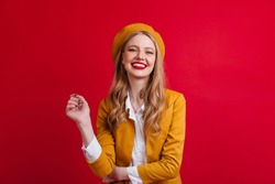Blissful french female model laughing at camera. Front view of blonde girl in beret isolated on red background.