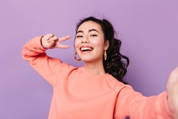 Lovely korean girl posing with peace sign. Laughing asian young woman taking selfie on purple background.