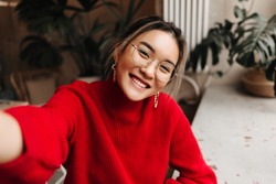 Lady in glasses and massive earrings makes selfie. Asian girl dressed in red knitted sweater posing against background of plants in room