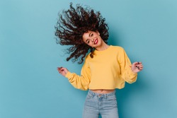 Brunette girl dances and plays wavy hair against blue background. Portrait of cute girl in yellow sweatshirt