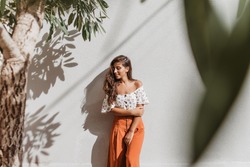 Attractive long-haired brunette woman in stylish culottes and white top posing against white wall and olive tree