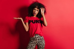 Wonderful young woman in t-shirt expressing unpleased emotions. Angry african girl posing on red background.