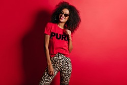 Joyful gorgeous woman in t-shirt smiling on red background. Indoor photo of african fascinating girl in sunglasses.