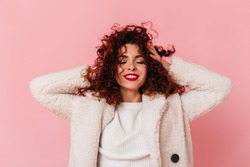 Portrait of charming lady with red lipstick and snow-white smile dressed in bright eco-coat and touching her curly hair on pink background