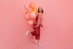 Full length view of joyful lady jumping with air balloons. Studio shot of laughing birthday girl posing on pink background.