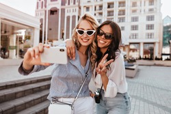 Well-dressed blonde lady making selfie with sister. Outdoor shot of romantic girls having fun in sunny spring day.
