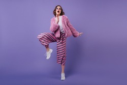 Funny european cheerful girl in white sneakers posing on purple background. Debonair young lady in striped pants dancing in studio.