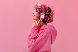 Profile portrait of charming pink-haired woman in massive headphones listening to music on isolated background. Curly girl in hoodie smiles.