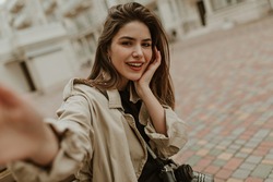 Young brunette woman in beige trench coat smiles widely and takes selfie outside. Attractive lady sits on wooden bench.