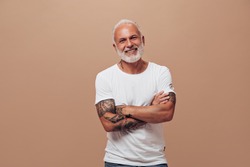 Grey haired man in white T-shirt poses on beige background. Handsome guy with beard in tattoos in bright clothes have fun into camera