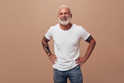 Tattooed man in good mood posing on beige background. Gray-haired guy in white T-shirt and blue jeans laughs into camera