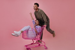 Positive man and woman having fun and riding shopping cart on pink background. Cheerful guy in brown sweater and pants posing with girlfriend on isolated