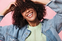 Smiling curly woman in denim jacket posing over pink background. Joyful happy girl in green tee laughing on isolated backdrop