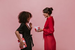 Trendy brunette lady with silver round earrings in red bright dress giving gift her curly friend in dark clothes with box.