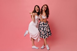 Brunette lady shows peace sign and poses with her friend on pink background. Girls with wavy hair in white fashionable dresses and converse hugging