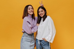 Cool joyful girl in purple sweater gives bunny ears to her smiling friend. Asian women in good mood pose on isolated orange background.