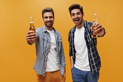 Excited young men in white trendy t-shirts and checkered shirts rejoice and hold beer bottles on orange isolated background.