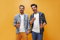 Joyful young brunet men in colorful shorts and checkered shirts laugh, point at camera and hold beer bottles on orange background.
