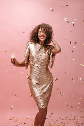 Cheerful girl with curly hairstyle in shiny trendy dress holding glass with drink and smiling with closed eyes on isolated backdrop..